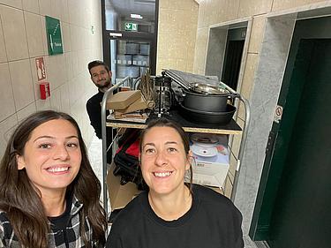 Fairschenkladen Drei Mitarbeitende der Eckert Schulen bringen neue Dinge in den Fairschenkladen. Sie stehen mit einem vollgepackten Wagen vor dem Aufzug. Es ist ein Selfie und alle sehen glücklich aus. Vorne stehen zwei Frauen hinten streckt ein Mann noch seinen Kopf hervor.