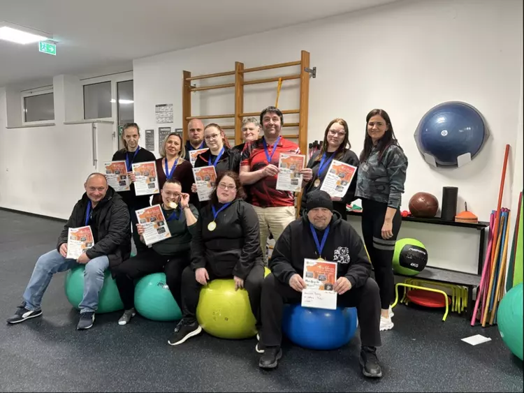 Gruppe von Teilnehmenden eines Gesundheitskurses sitzt und steht in einem Trainingsraum auf Gymnastikbällen und hält Urkunden sowie Medaillen in die Kamera.