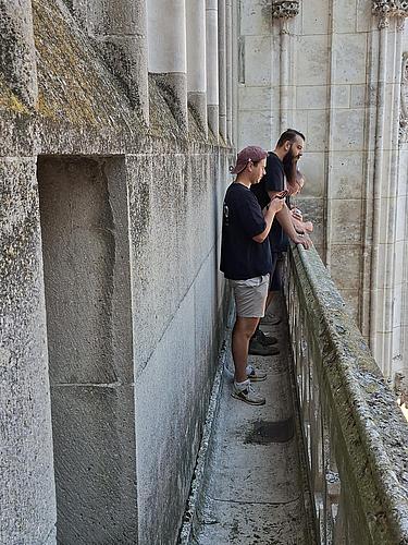 Drei Männer stehen auf einem sehr schmalen Steinumgang entlang der Fassade des Regensburger Doms. Sie lehnen sich an das steinerne Geländer und blicken nach unten. Die Fassade ist mit senkrechten gotischen Strebepfeilern und verwittertem Stein versehen.