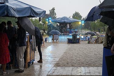 Eine Gruppe von Menschen steht bei Regen unter großen blauen Sonnenschirmen und Pavillons an einem Strandabschnitt mit Sandboden. Im Hintergrund ist eine beleuchtete Musikbühne sichtbar.